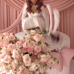 A woman wearing Ivory Sheep's Eloise Mini Lace Dress is sitting on a large cake with pink flowers and ribbons against a pink curtain background.