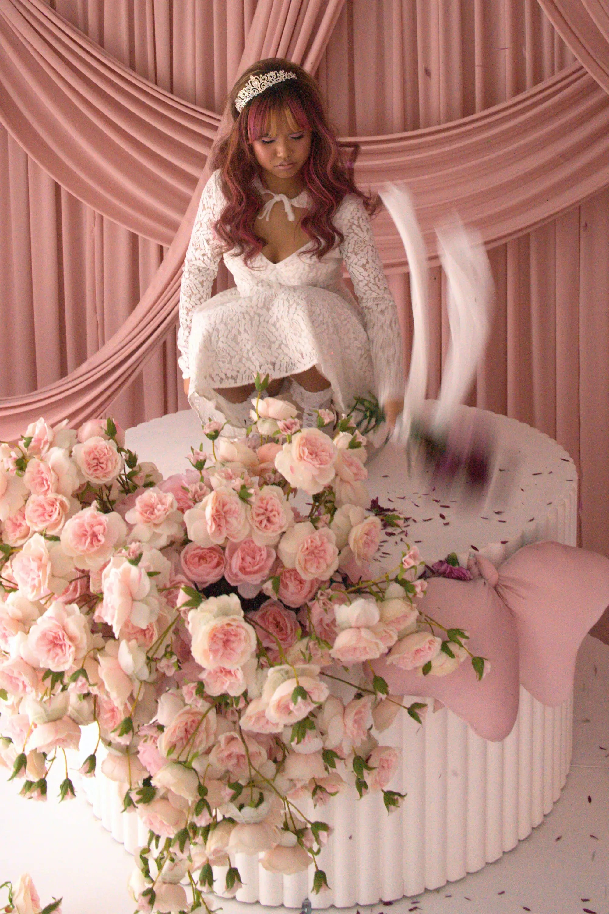 A woman wearing Ivory Sheep's Eloise Mini Lace Dress is sitting on a large cake with pink flowers and ribbons against a pink curtain background.