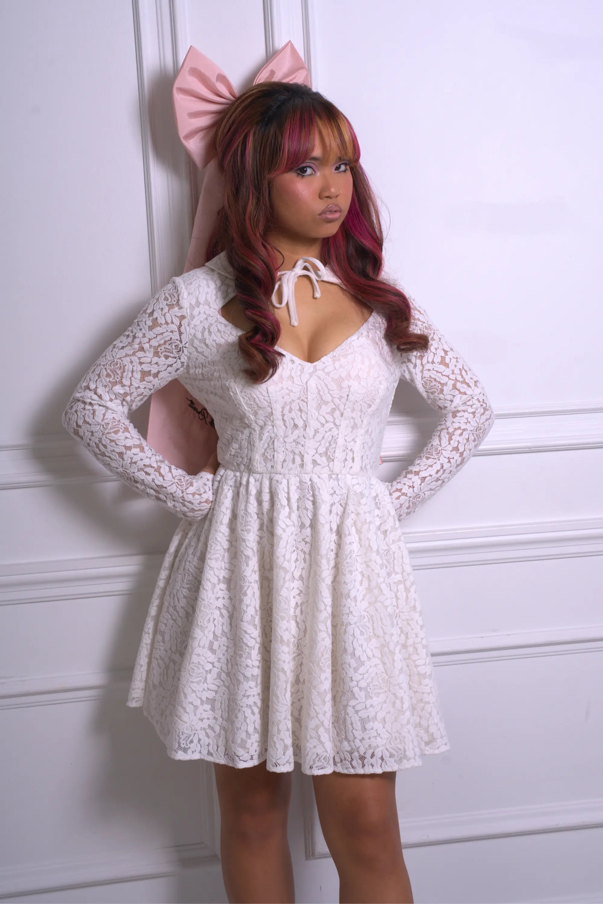 A woman wearing Ivory Sheep's Eloise Mini Lace Dress is standing against a white wall.
