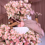 A woman wearing Ivory Sheep's Garden Party Lace Midi Dress lying on a bed of pink flowers with a pink curtain backdrop
