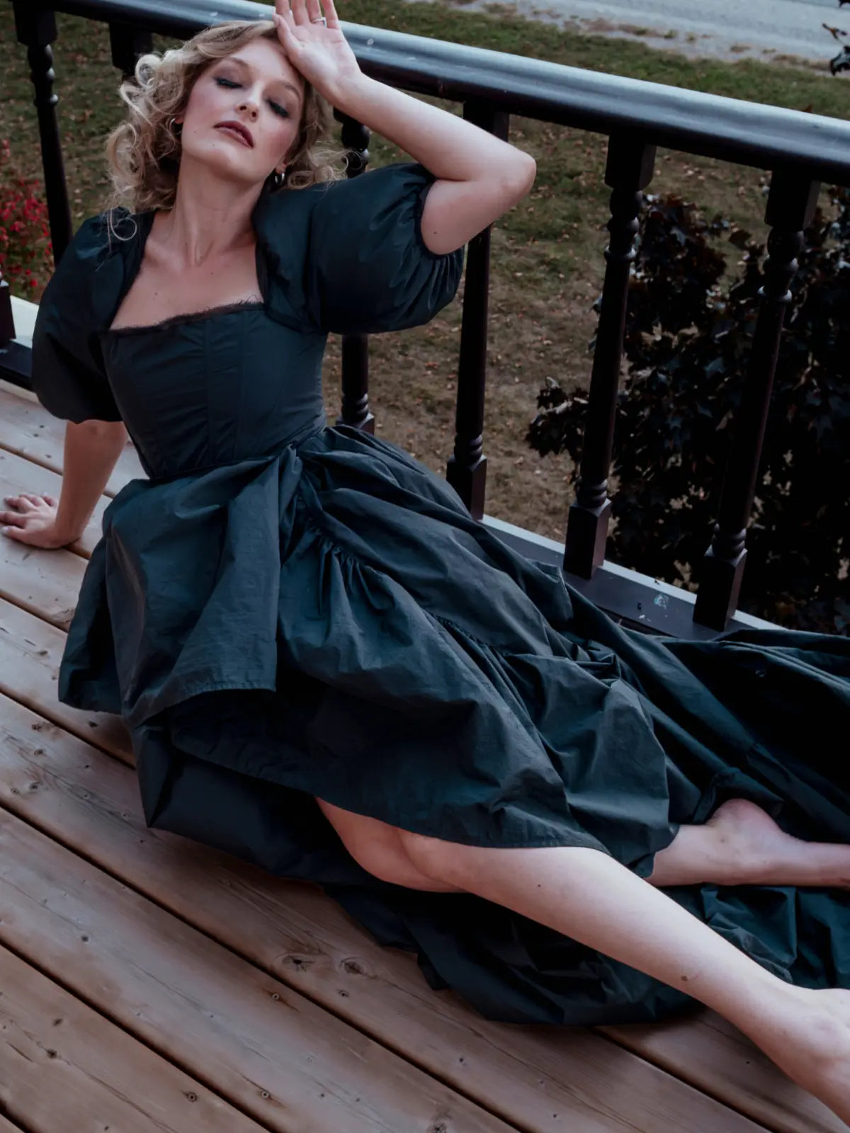 A woman in a black taffeta corset dress sitting on a wooden deck with a blurred background.