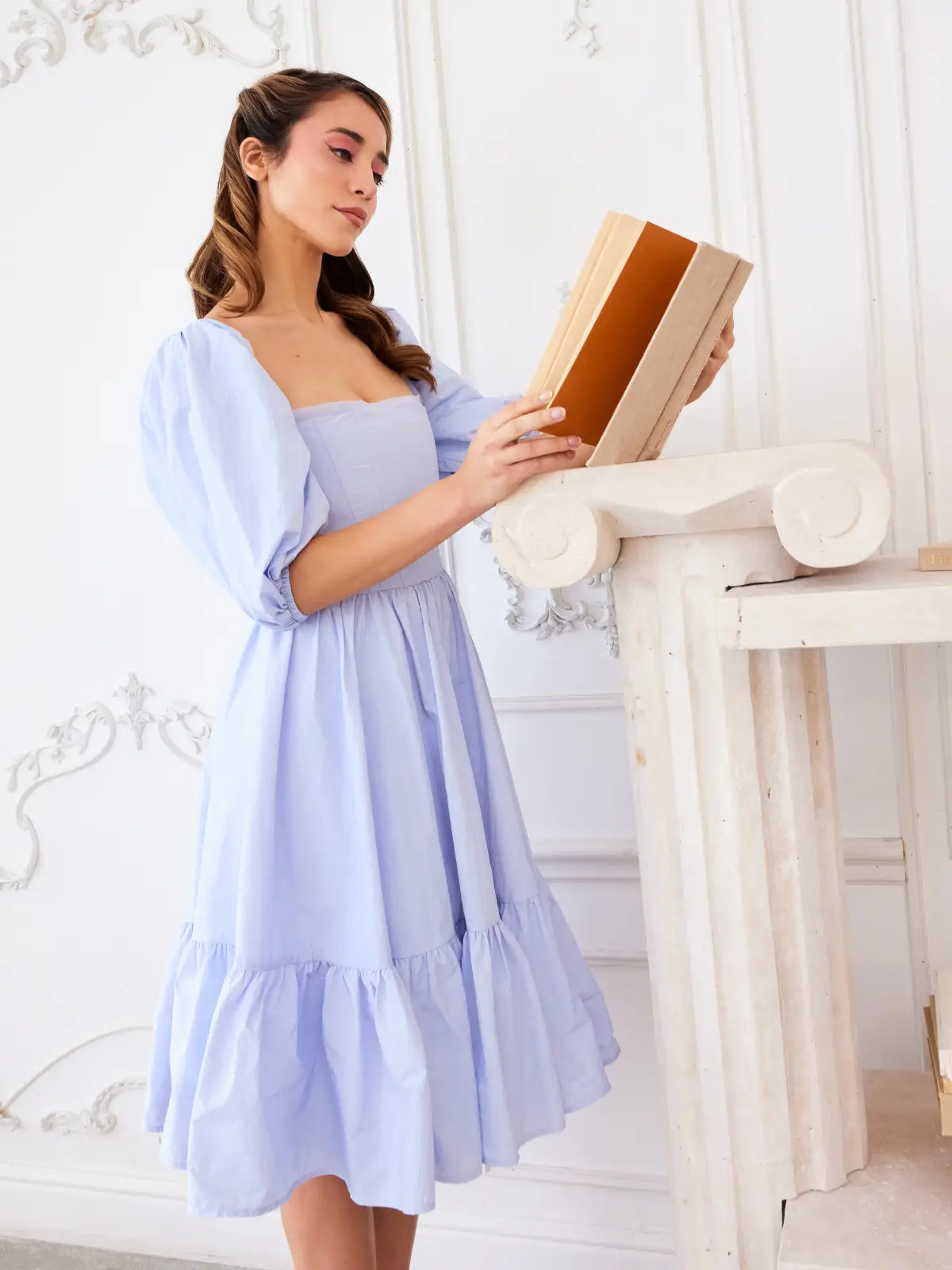 A woman wearing the Alice blue corset mini dress holding a book in a bright room with white walls and furniture.