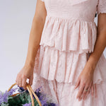 A woman wearing Ivory Sheep's Leighton Pink Cotton Mini Dress holding a basket of flowers against a white background.