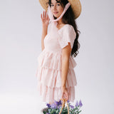 A woman wearing Ivory Sheep's Leighton Pink Cotton Mini Dress and straw hat holding a basket of flowers against a white background