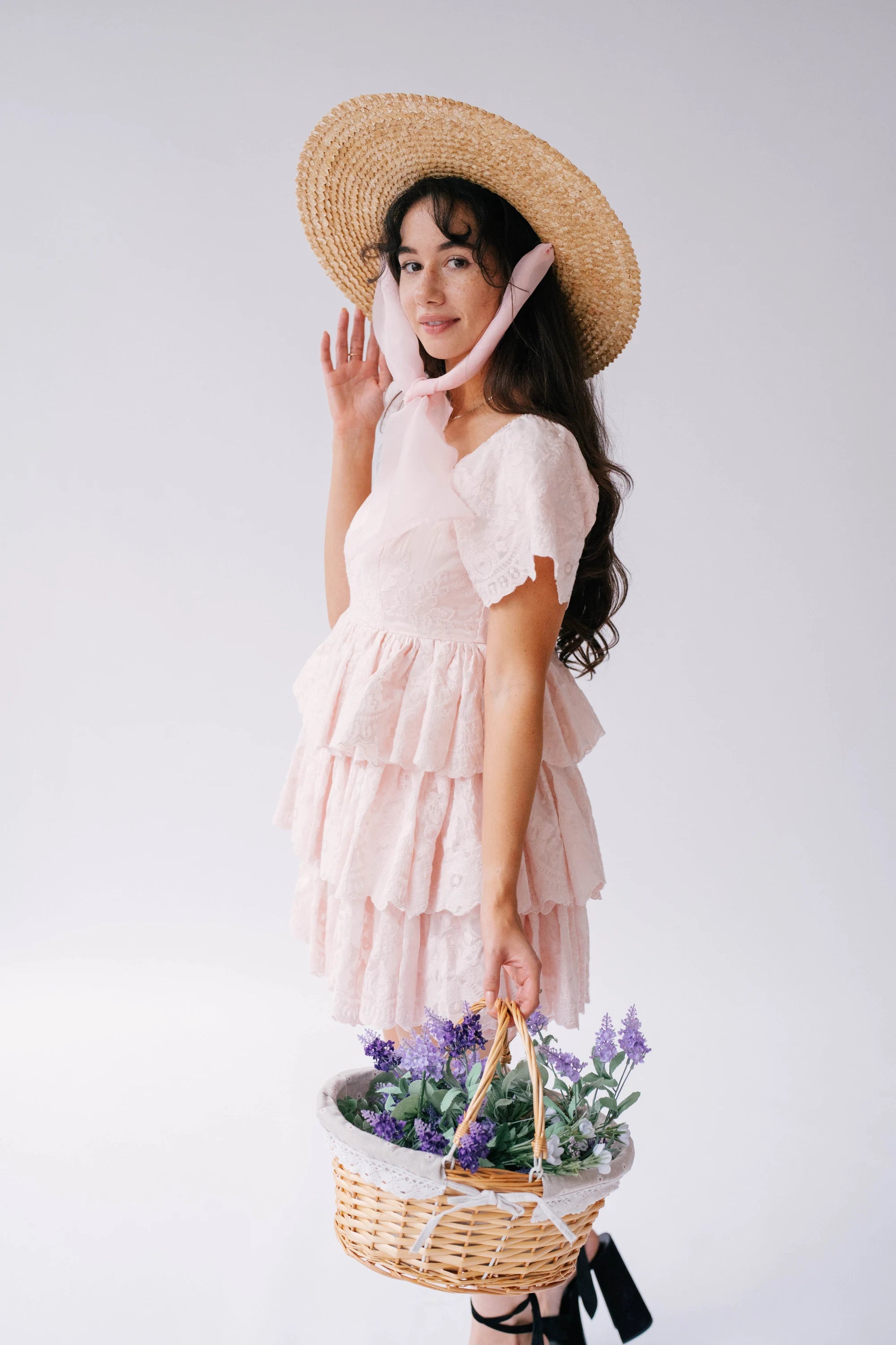 A woman wearing Ivory Sheep's Leighton Pink Cotton Mini Dress and straw hat holding a basket of flowers against a white background