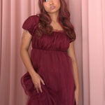 A woman wearing Ivory Sheep's crimson red Molly Midi Dress with puffed sleeves against a pink curtain background