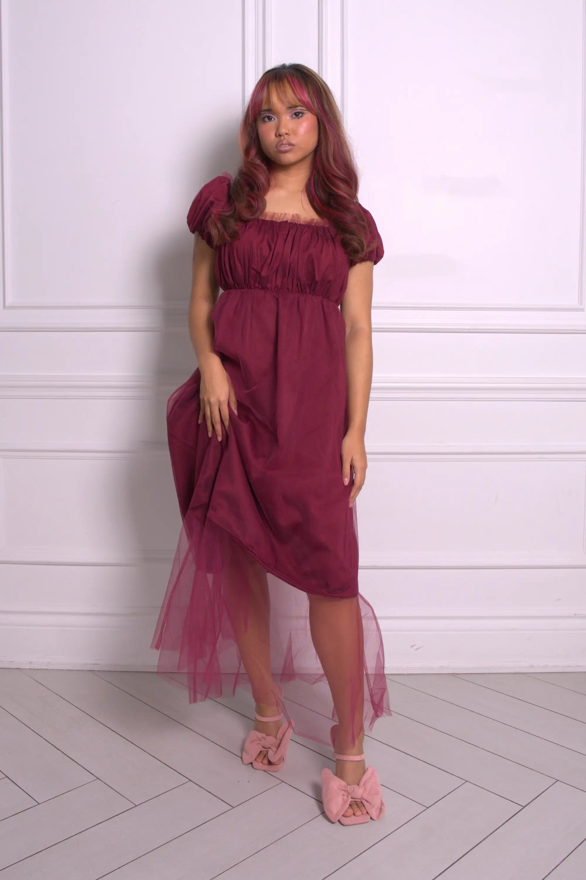 A woman wearing Ivory Sheep's crimson red Molly Midi Dress with a sheer overlay against a white paneled wall.