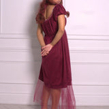 A woman wearing Ivory Sheep's crimson red Molly Midi Dress with sheer overlay against a white paneled wall.