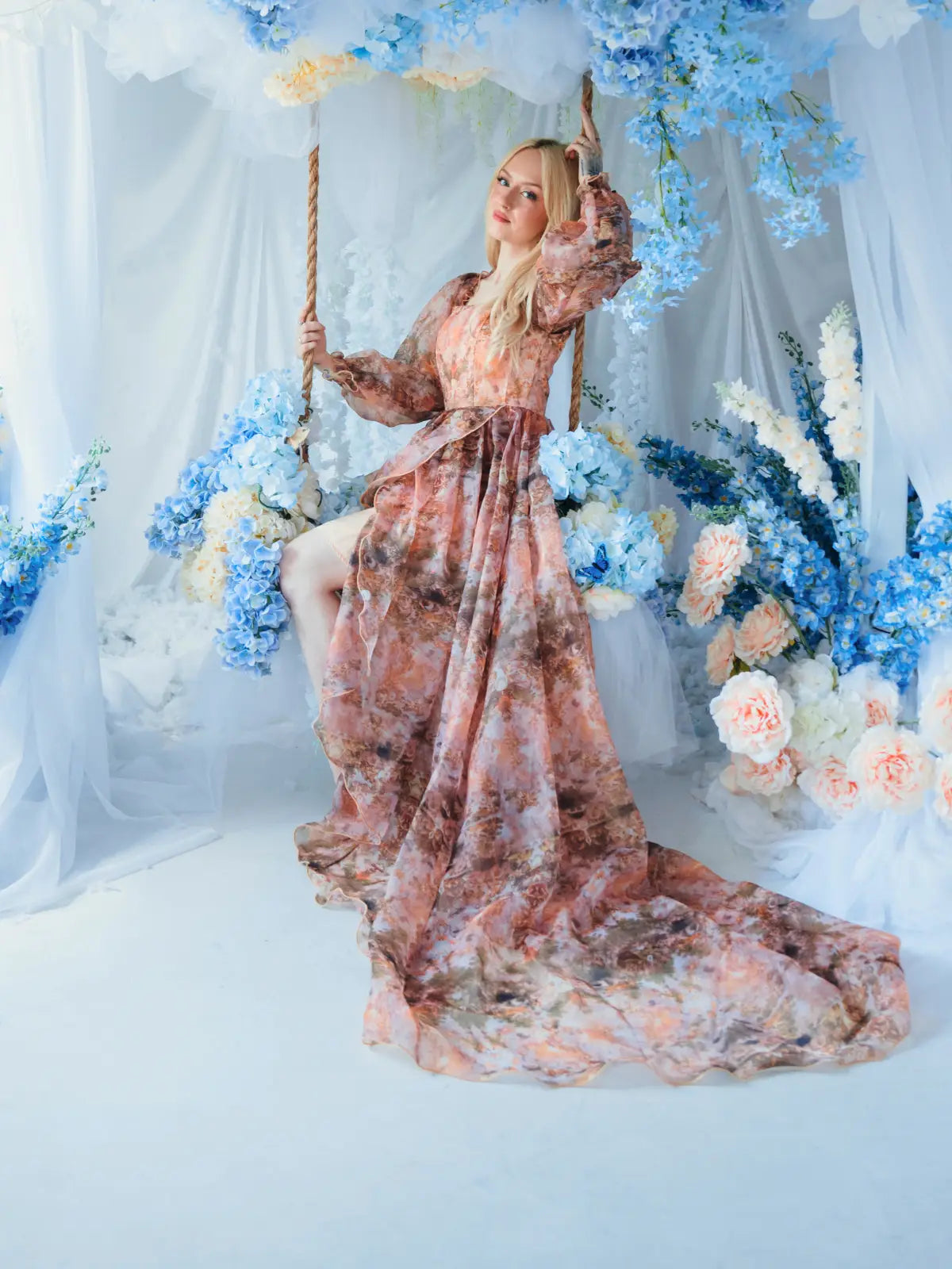 A woman in Ivory Sheep's Ode to Jove Reverie Gown sitting on a swing surrounded by blue and white floral decorations.