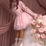 A woman wearing Ivory Sheep's Strawberry Riot Pink Lace Bolero is standing next to a floral arrangement against a pink curtain backdrop.
