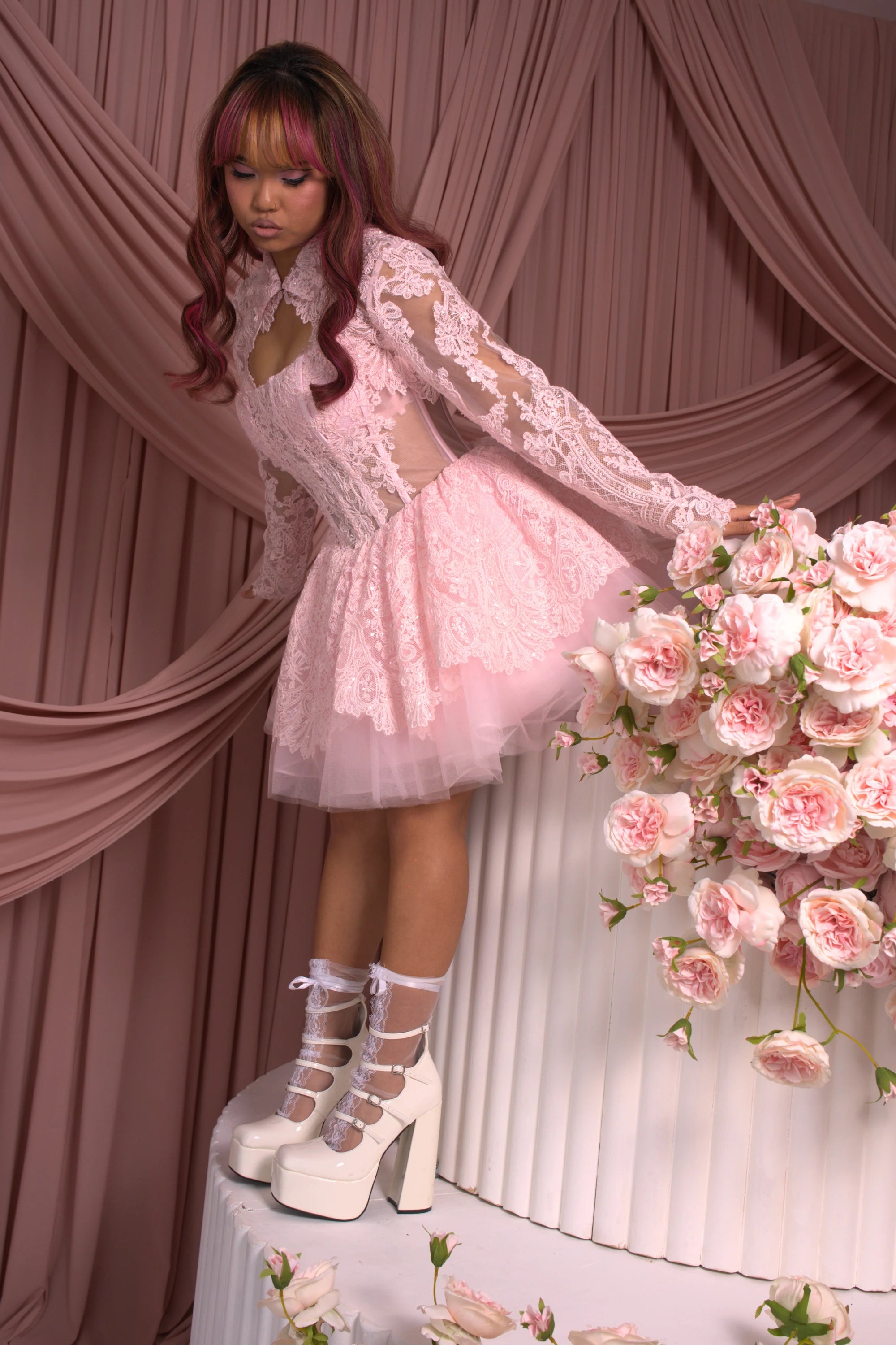 A woman wearing Ivory Sheep's Strawberry Riot Pink Lace Bolero is standing next to a floral arrangement against a pink curtain backdrop.