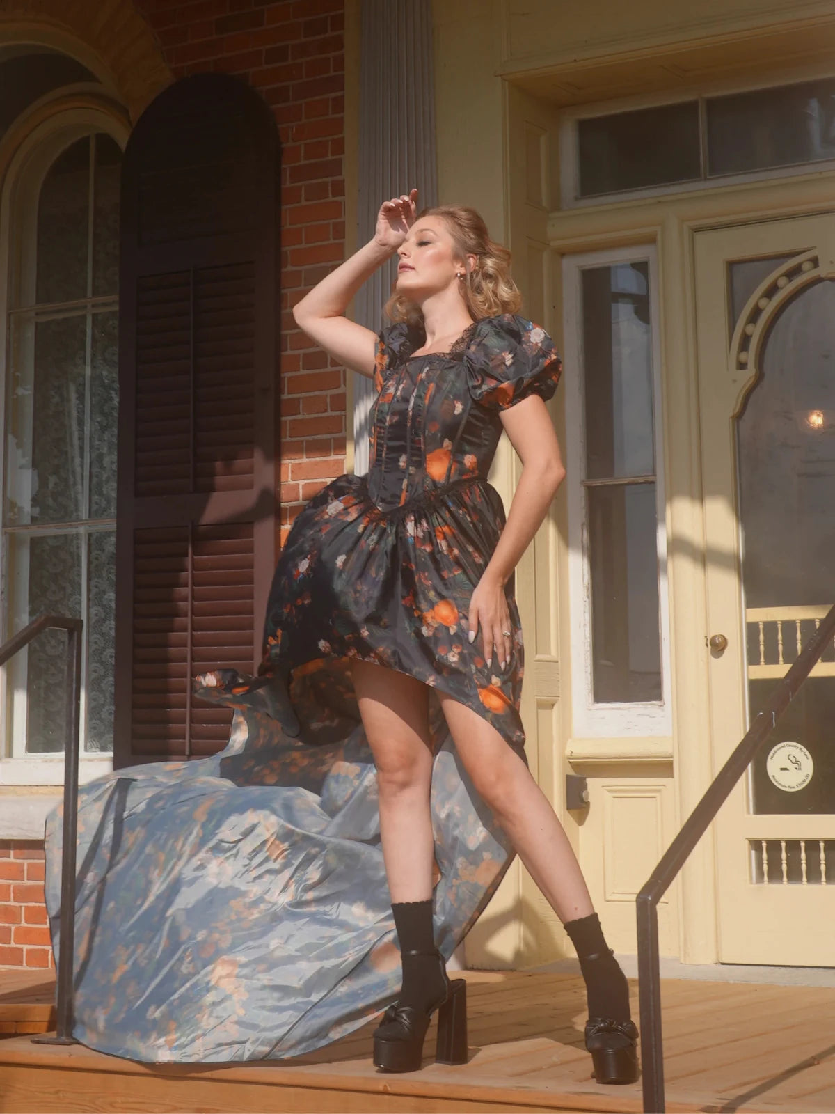 Woman in a floral dress standing on a porch with a vintage house in the background