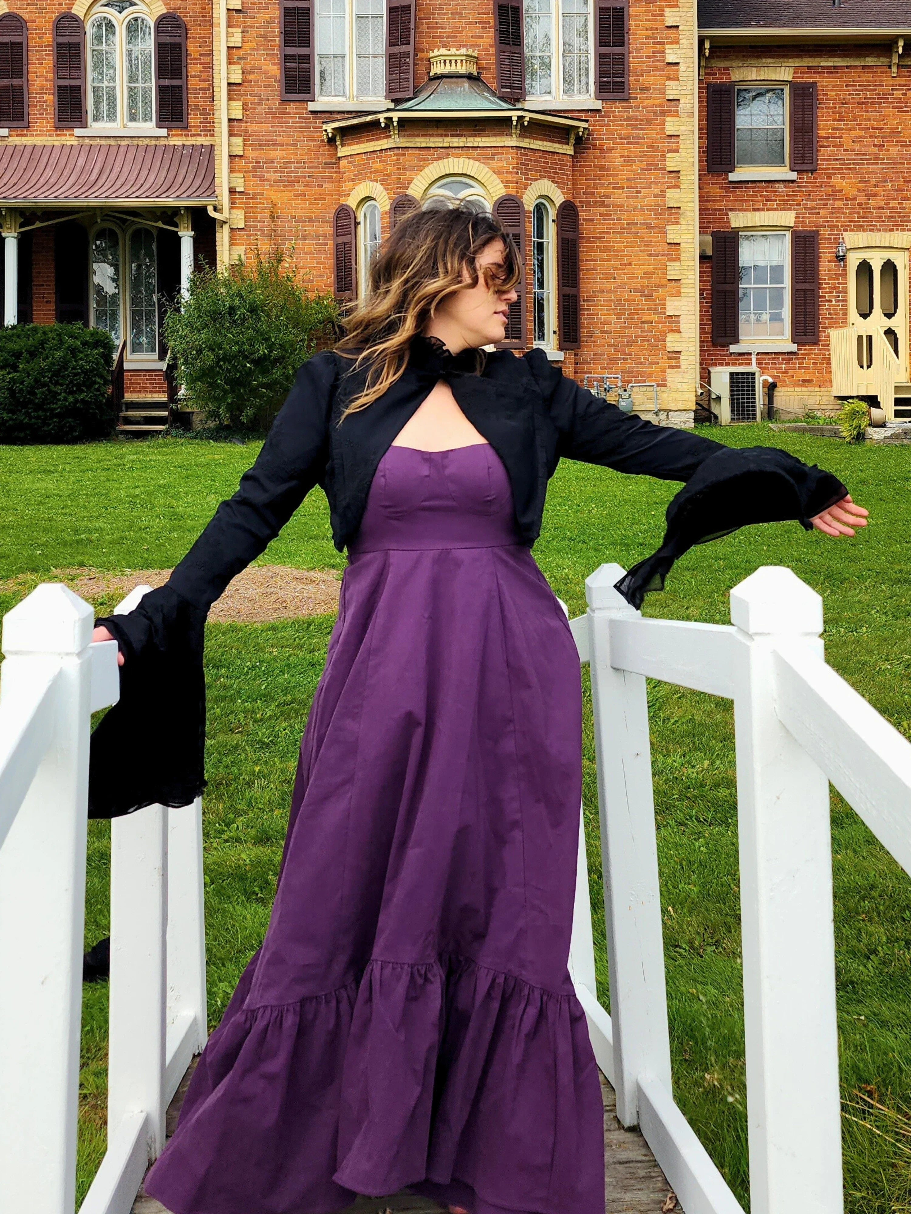 Woman in a purple dress and Lady Elizabeth jacket posing by a white fence