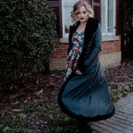 A woman in a long black coat and a floral dress is standing outside near a brick building.
