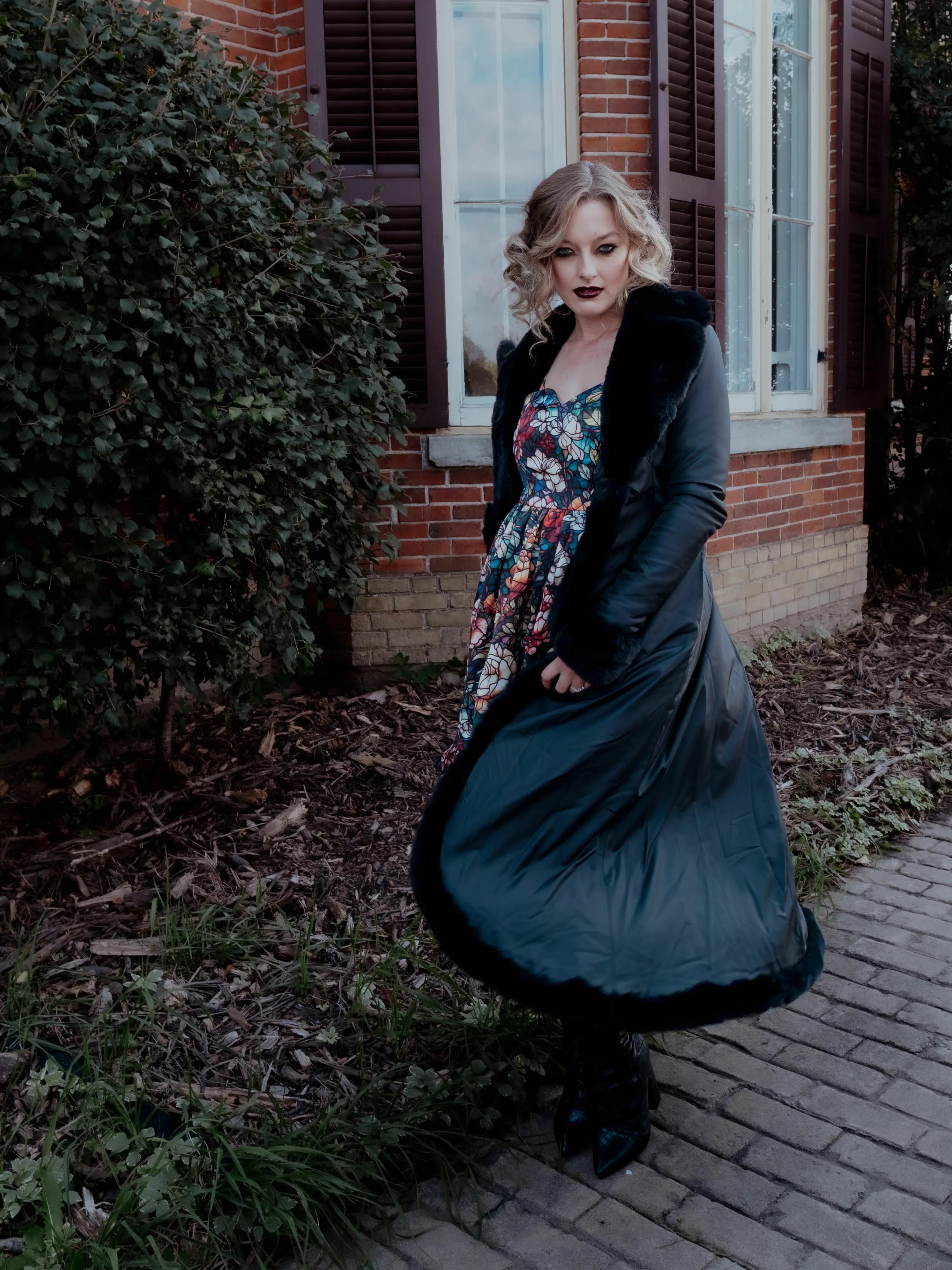 A woman in a long black coat and a floral dress is standing outside near a brick building.