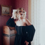 A woman in a black off-the-shoulder mini dress is standing in a room with antique red walls and an old painting.