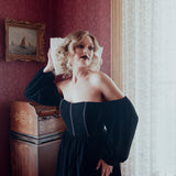 A woman in a black off-the-shoulder mini dress is standing in a room with antique red walls and an old painting.