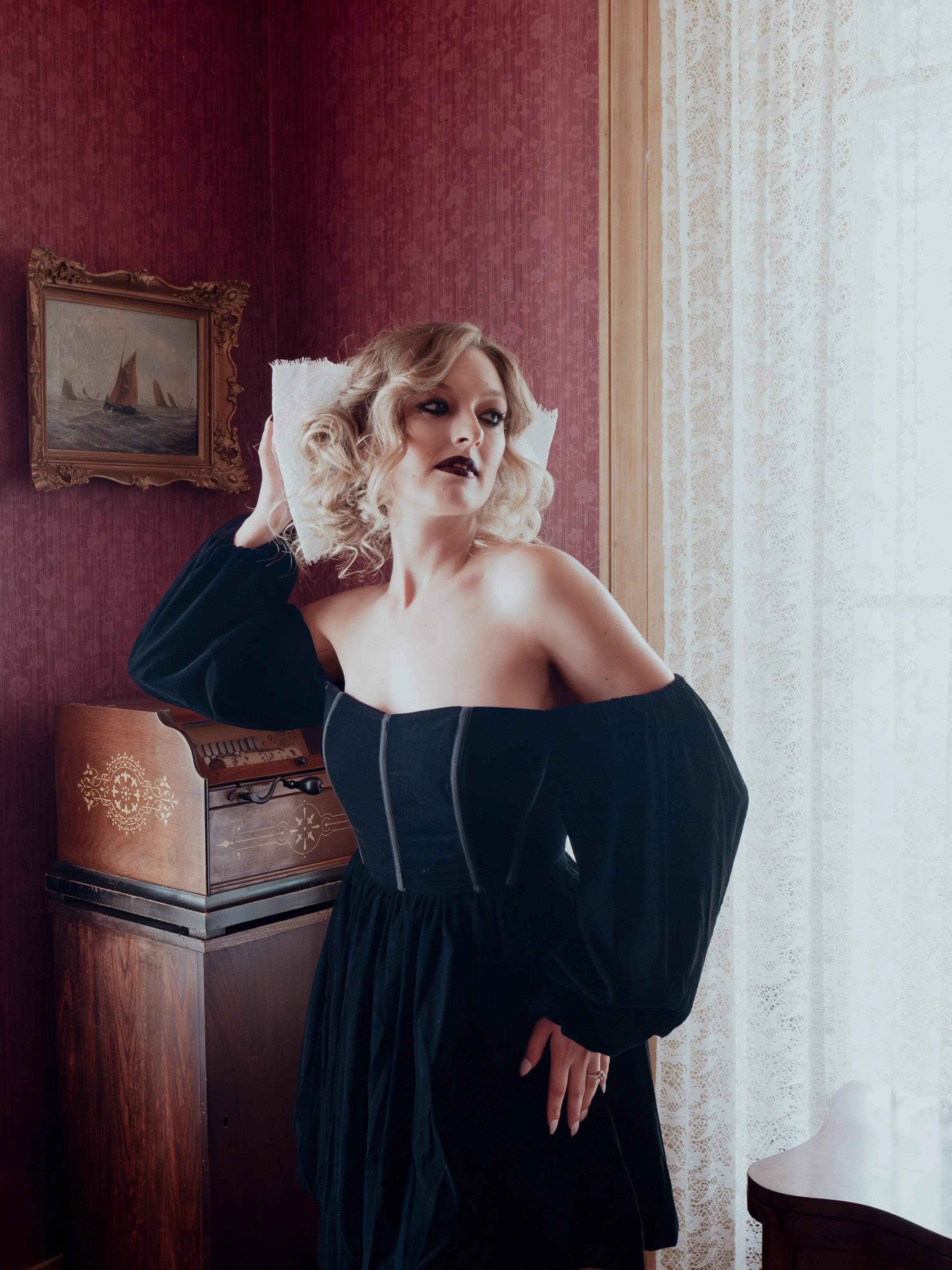 A woman in a black off-the-shoulder mini dress is standing in a room with antique red walls and an old painting.