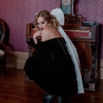 A woman crouched down wearing the Nocturne Velvet Mini Dress with a white hair bow in an antique room with purple walls and wooden furniture.