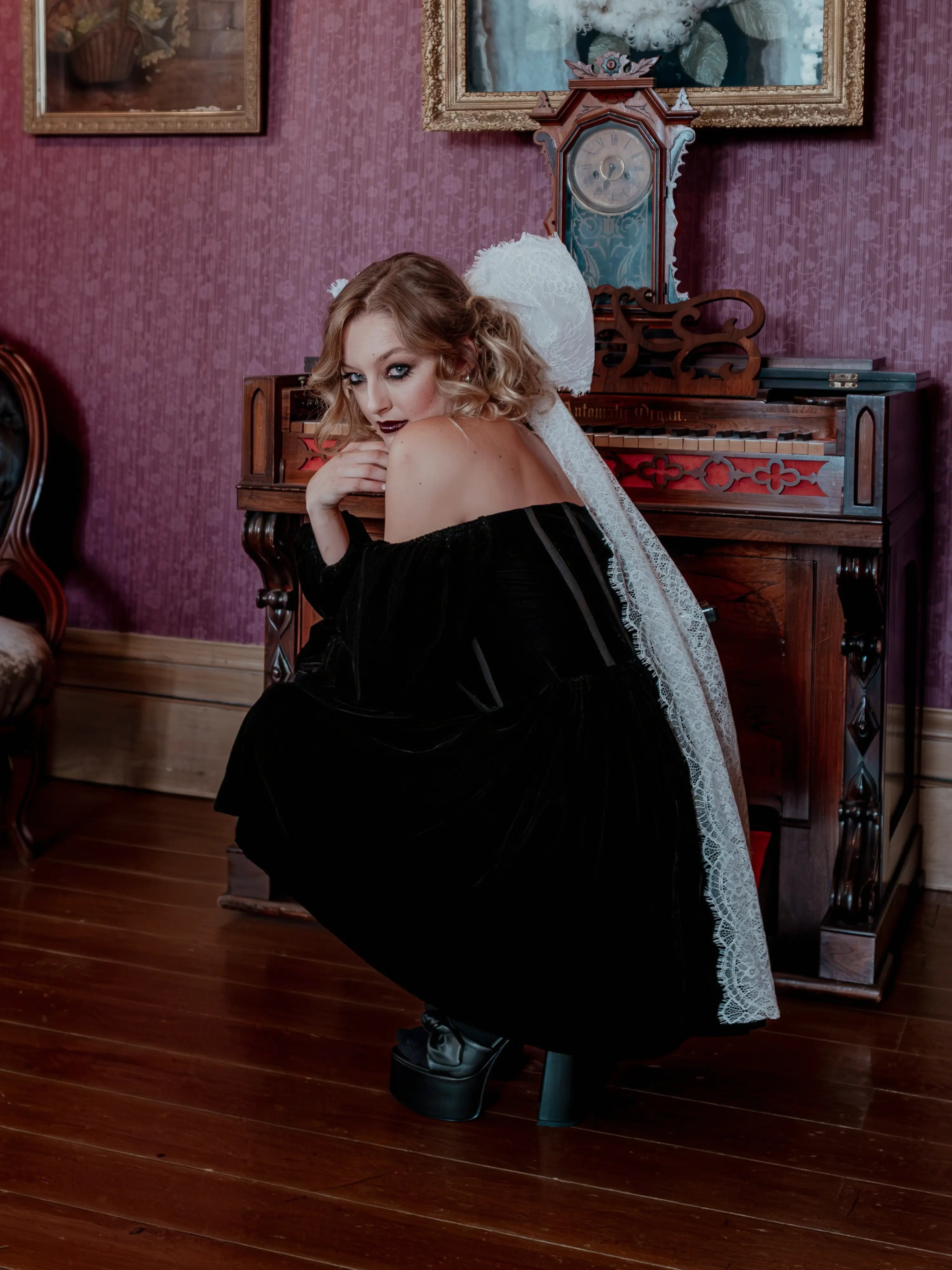 A woman crouched down wearing the Nocturne Velvet Mini Dress with a white hair bow in an antique room with purple walls and wooden furniture.