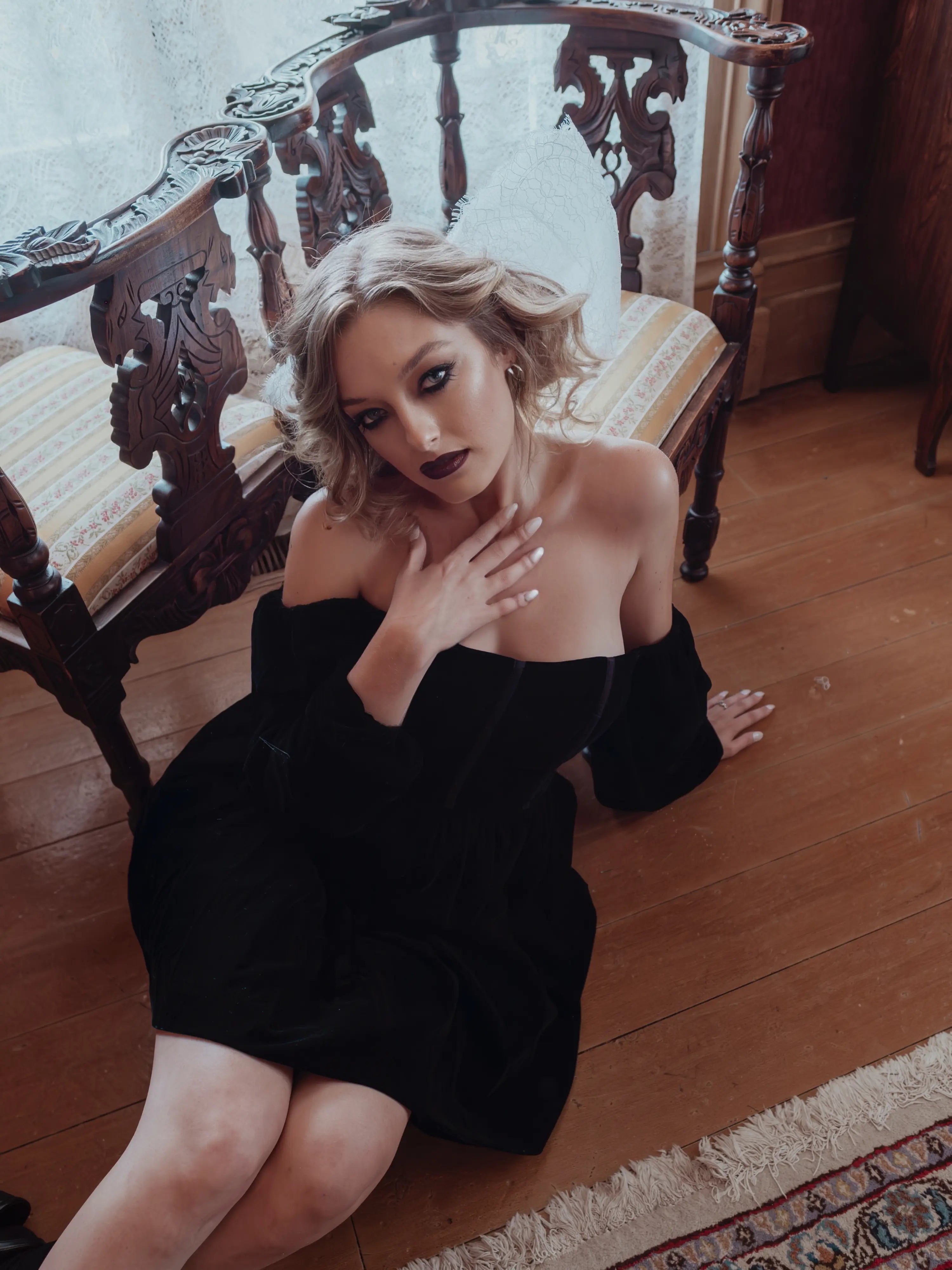 A woman in a black velvet mini dress dress sitting on a wooden floor with a decorative chair in the background.
