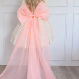 A full view photo of a woman wearing a white lace dress and large peach organza bow, showcasing the 10 foot long tails of the bow.