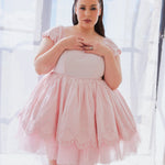 A full view of a woman wearing the blossom pink cotton mini dress in front of a window.