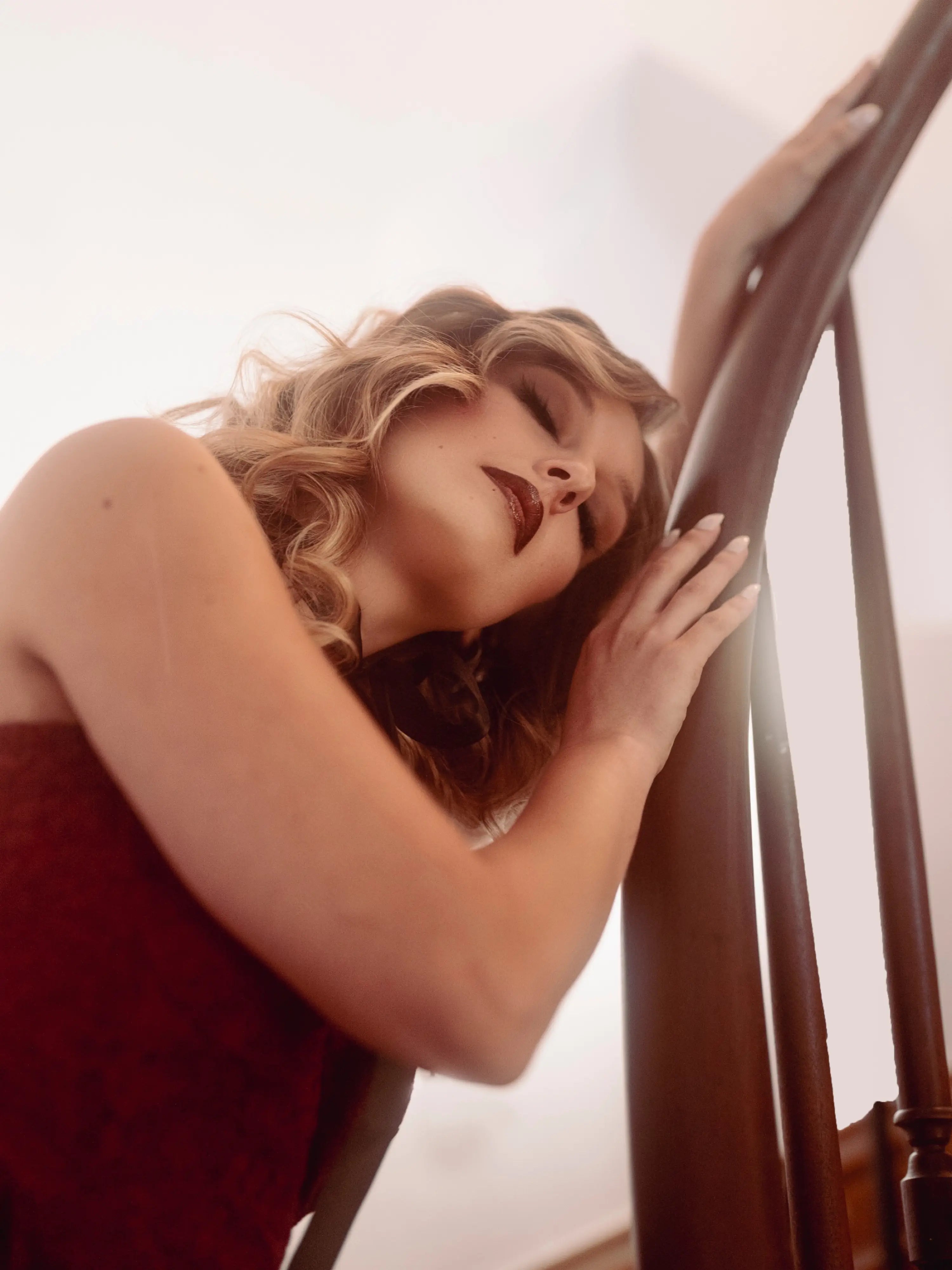 A woman in a red lace dress leaning against a wooden railing with a blurred background