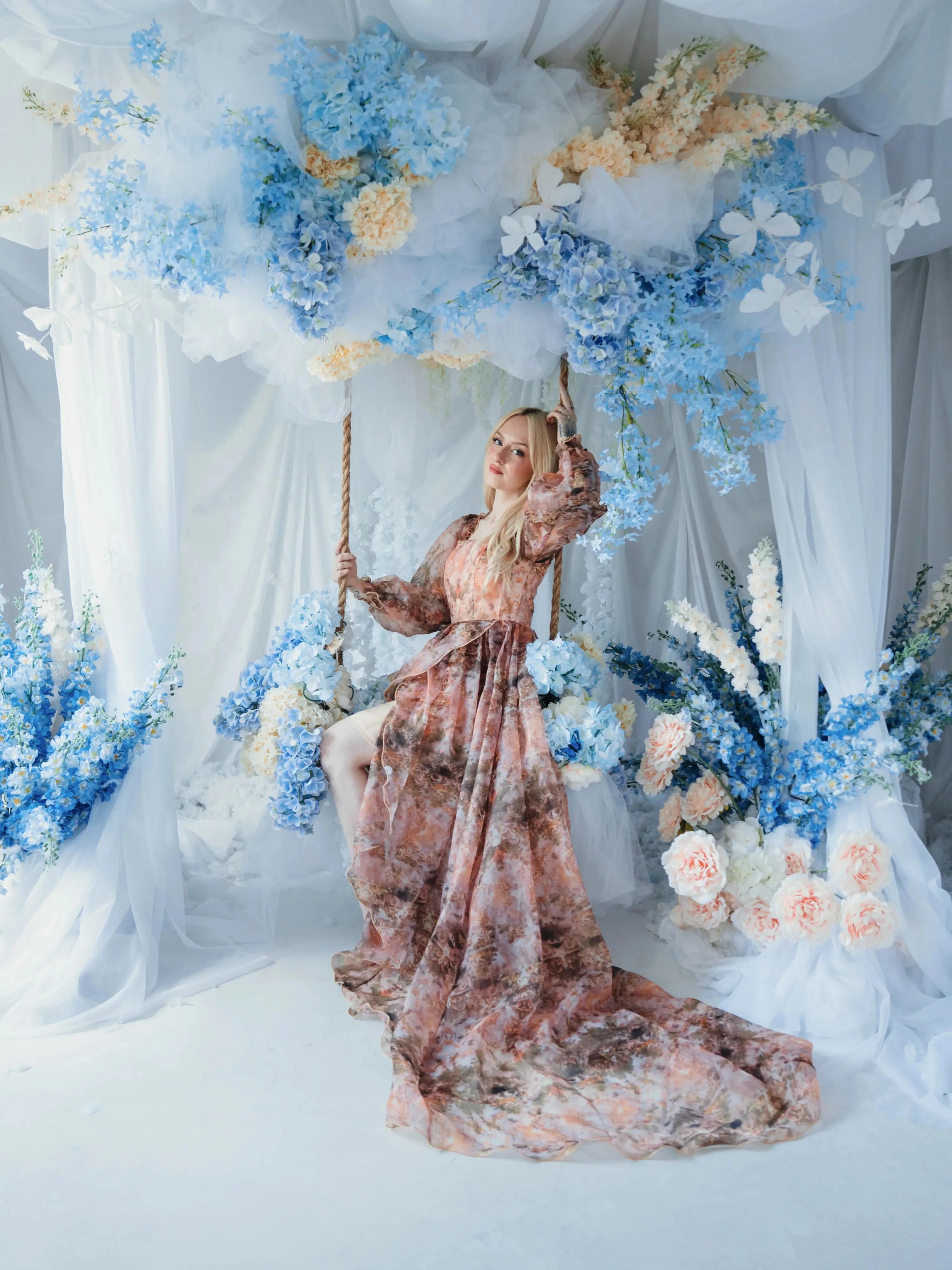 The Ode to Jove Twilight Reverie Ball Gown styled on a swing with trailing organza and floral backdrop, evoking a fairytale garden scene.