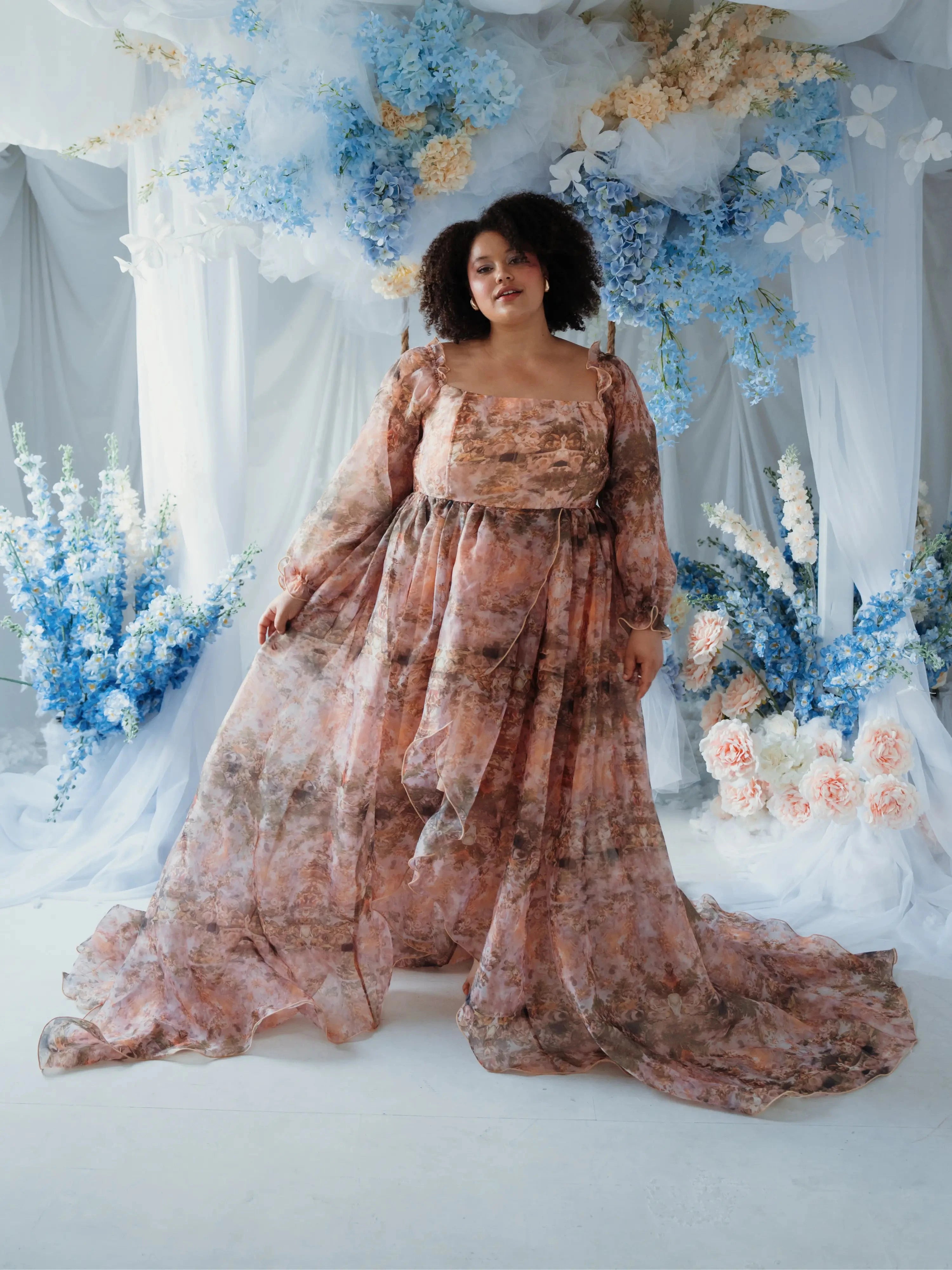 Plus-size model in the Ode to Jove Twilight Reverie Ball Gown with dramatic train and floral print, styled in a garden-inspired setting.