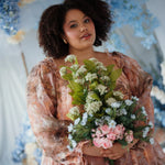 Plus sized woman in the Ode to Jove Twilight Reverie Ball Gown holding floral bouquet, surrounded by ethereal blue and white floral decor.