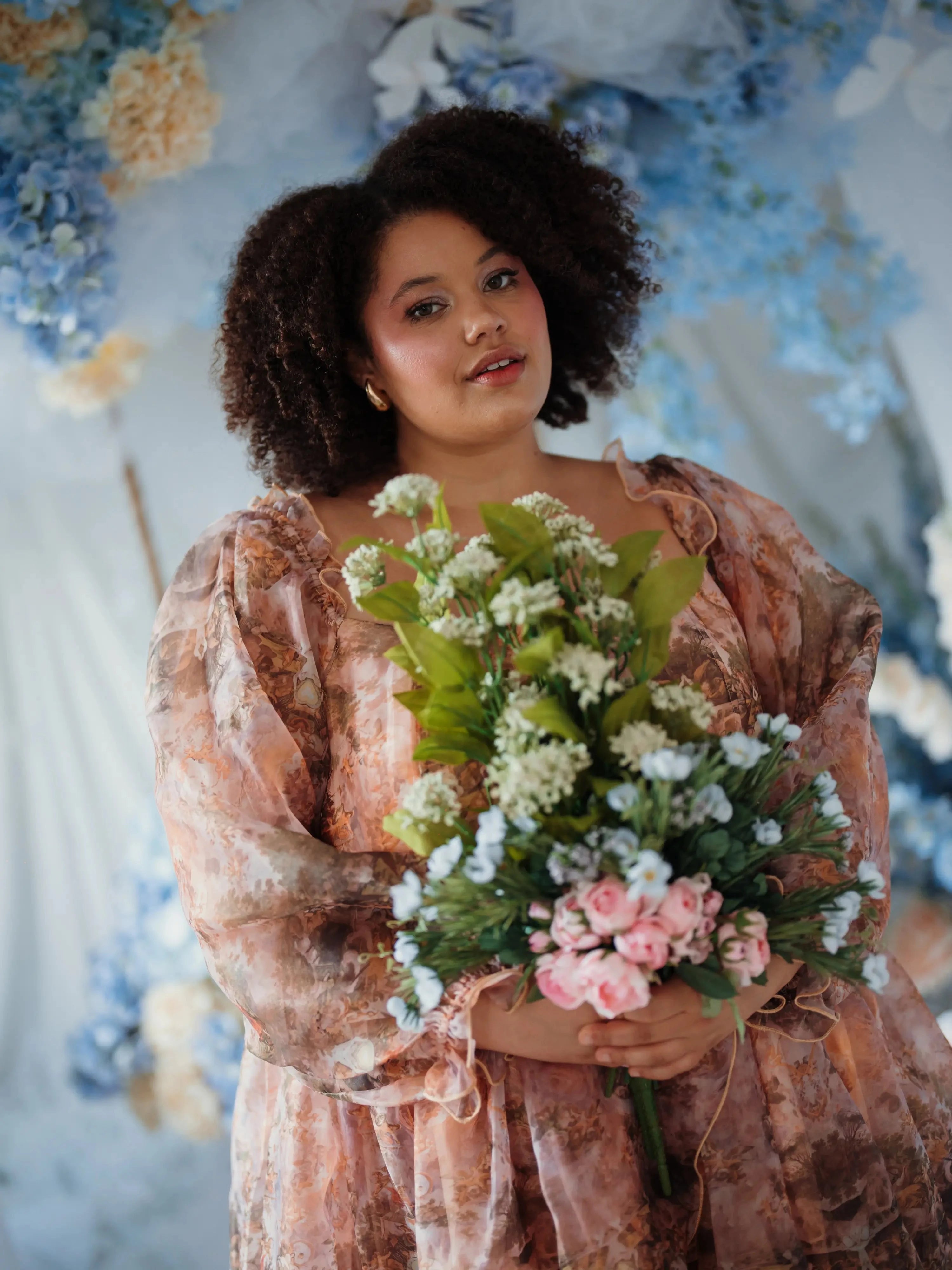 Plus sized woman in the Ode to Jove Twilight Reverie Ball Gown holding floral bouquet, surrounded by ethereal blue and white floral decor.