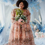 Plus-size woman in the Ode to Jove Twilight Reverie Ball Gown holding a bouquet, styled in a dreamy floral backdrop with blue and white flowers.