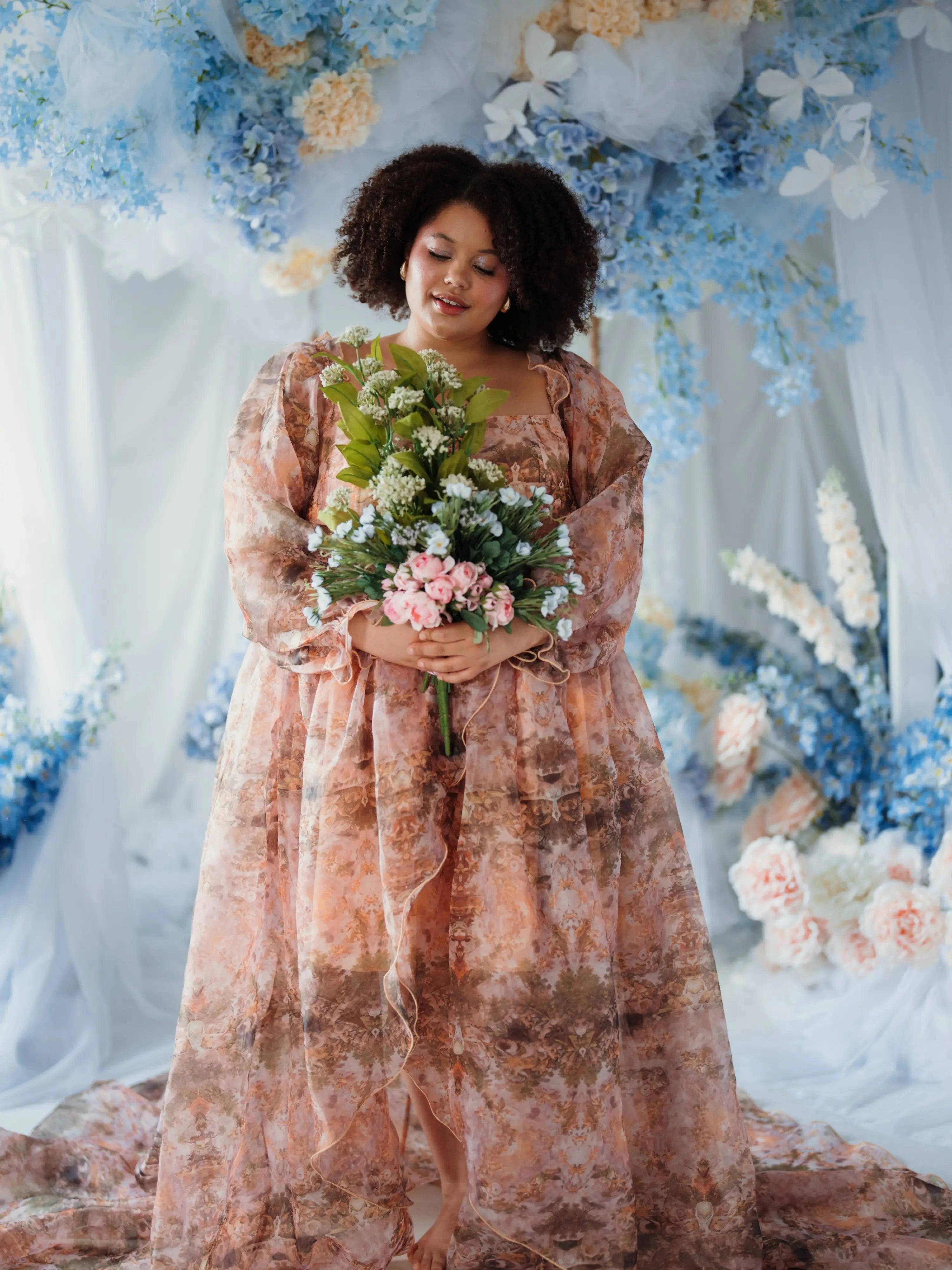 Plus-size woman in the Ode to Jove Twilight Reverie Ball Gown holding a bouquet, styled in a dreamy floral backdrop with blue and white flowers.