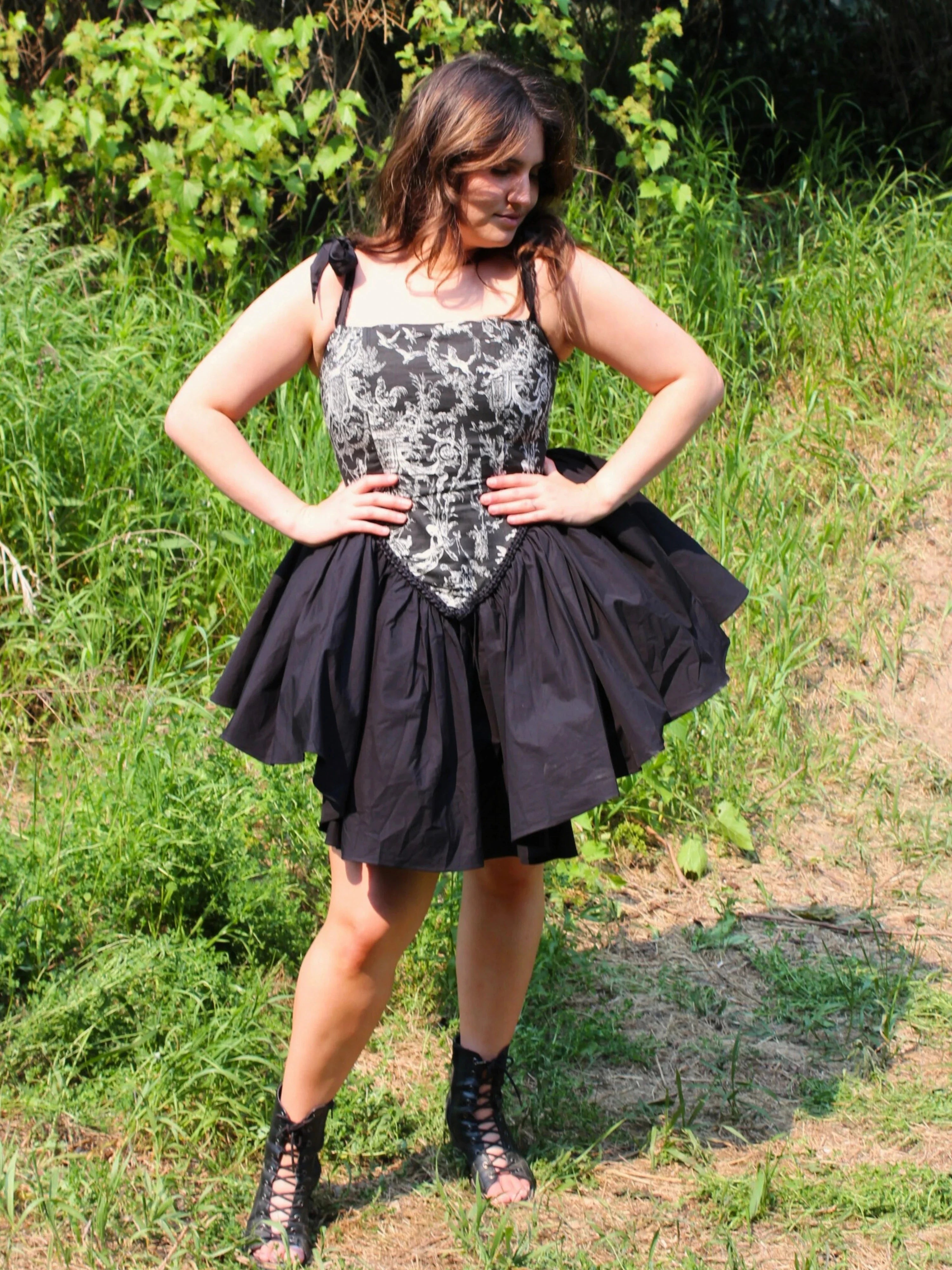A woman modeling the Rhiannon Corset Mini Dress with custom black toile print and lace-up sandals in a forest for a fairy tale look.
