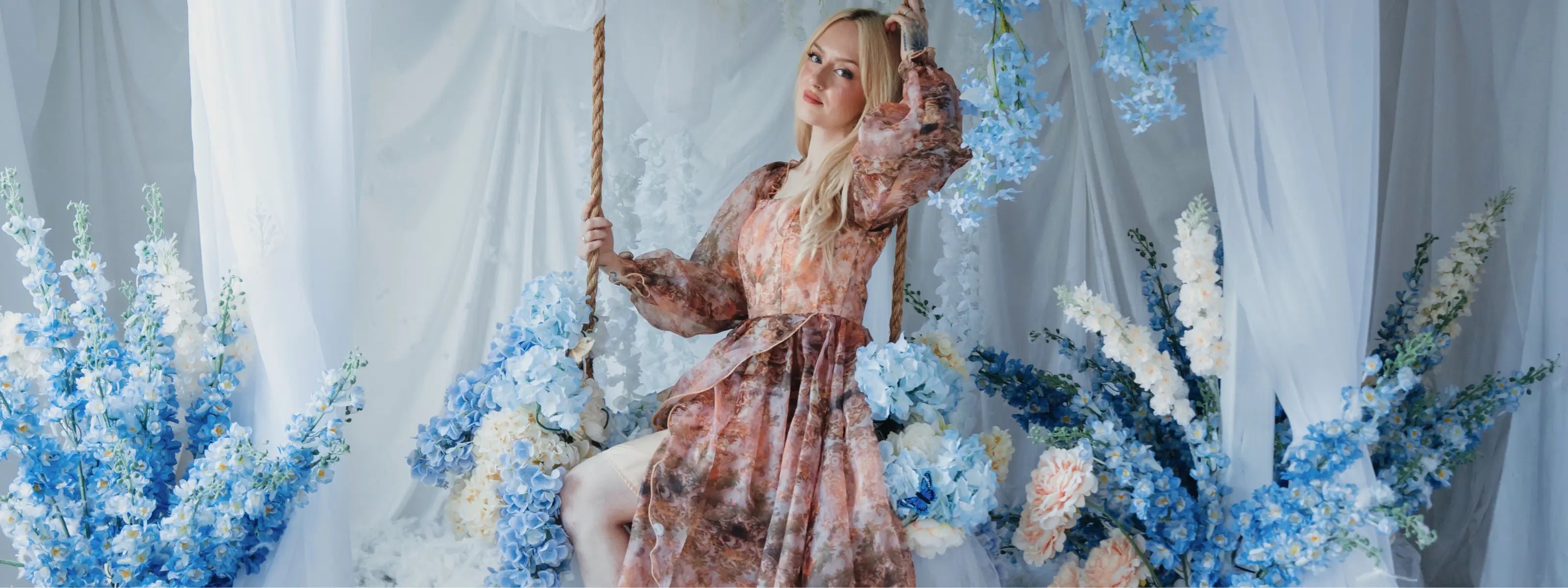 Woman in the ode to jove reverie ball gown sitting on a swing surrounded by blue flowers against a white curtain.