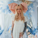 The Baby Antoinette Silk Hair Bow worn with a white lace dress, set against pastel floral decor for a coquette-inspired fashion look.