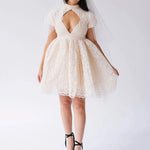 Woman wearing a white lace mini dress with a large diamond cutout on bust against a white background.