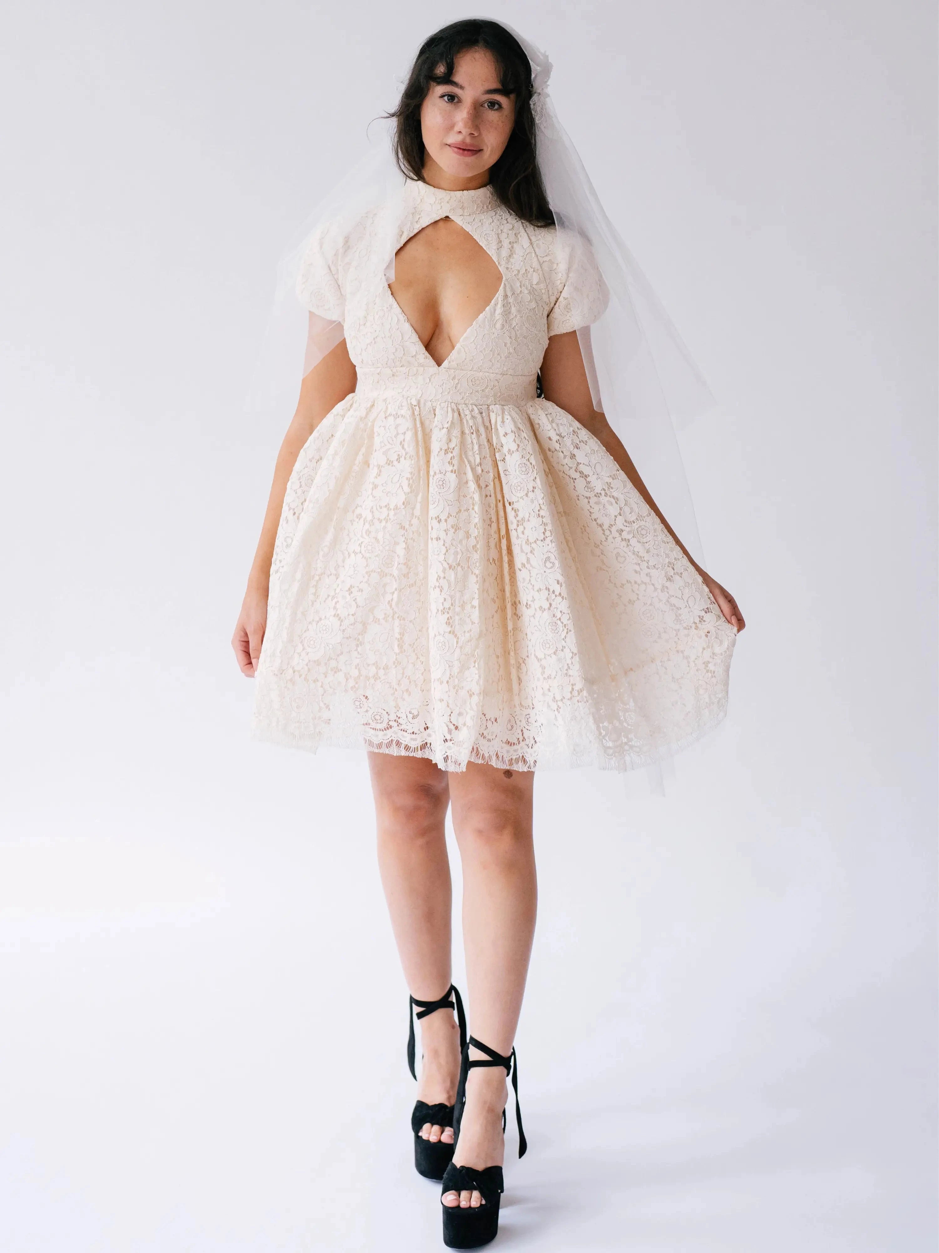 Woman wearing a white lace mini dress with a large diamond cutout on bust against a white background.