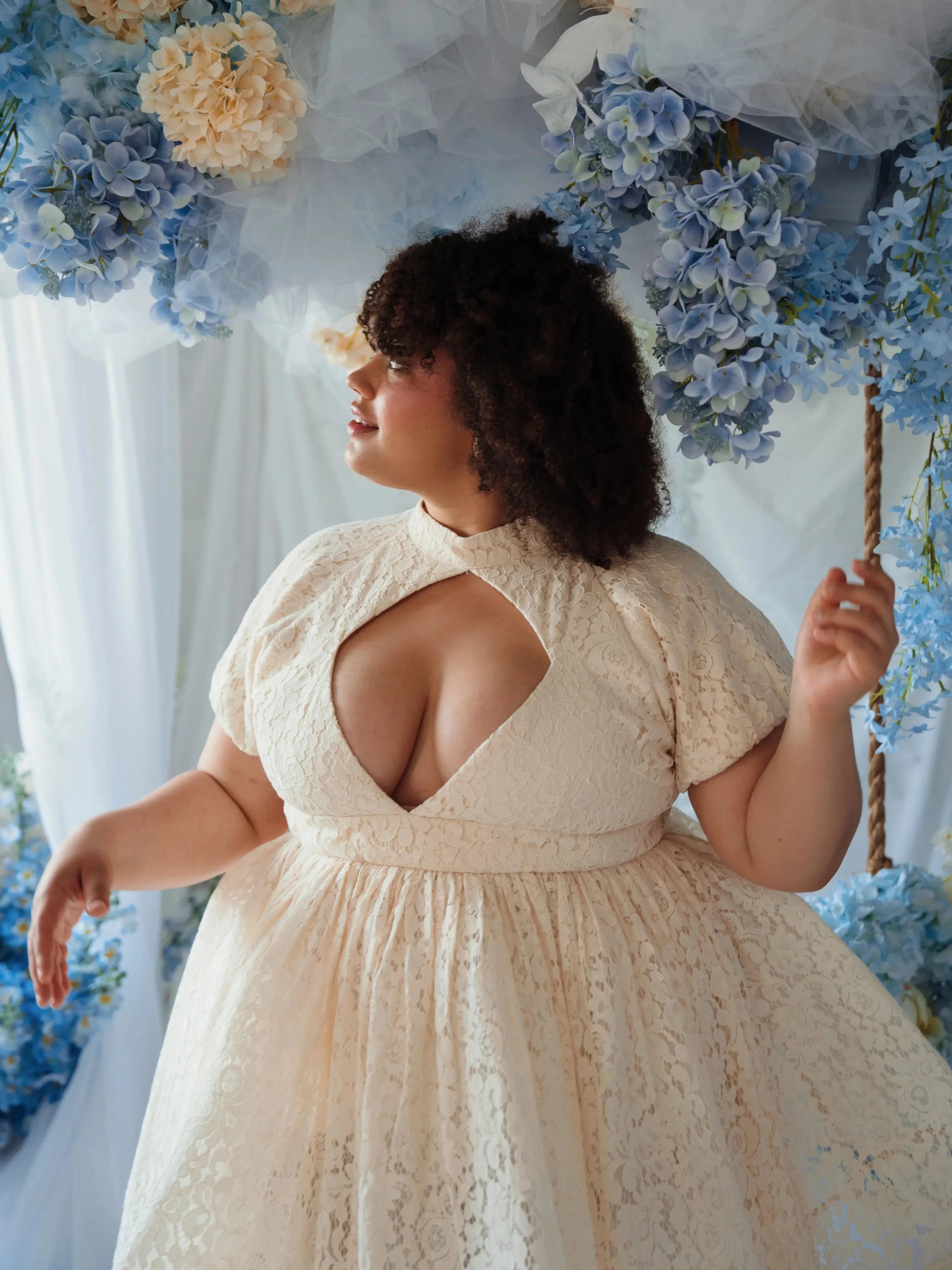 Plus-size model wearing The Victorian Lace Tea Party Mini Dress with diamond keyhole bust and high waist, surrounded by pastel hydrangeas.