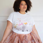 The Little Lamb Tee paired with a peach floral skirt, showcasing lambs, wildflowers, and butterflies in a soft cottagecore aesthetic.