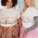 The Little Lamb Tee styled with pastel skirts, featuring whimsical lamb and floral artwork by Kaeti MacNeil. Cottagecore fashion for spring looks.