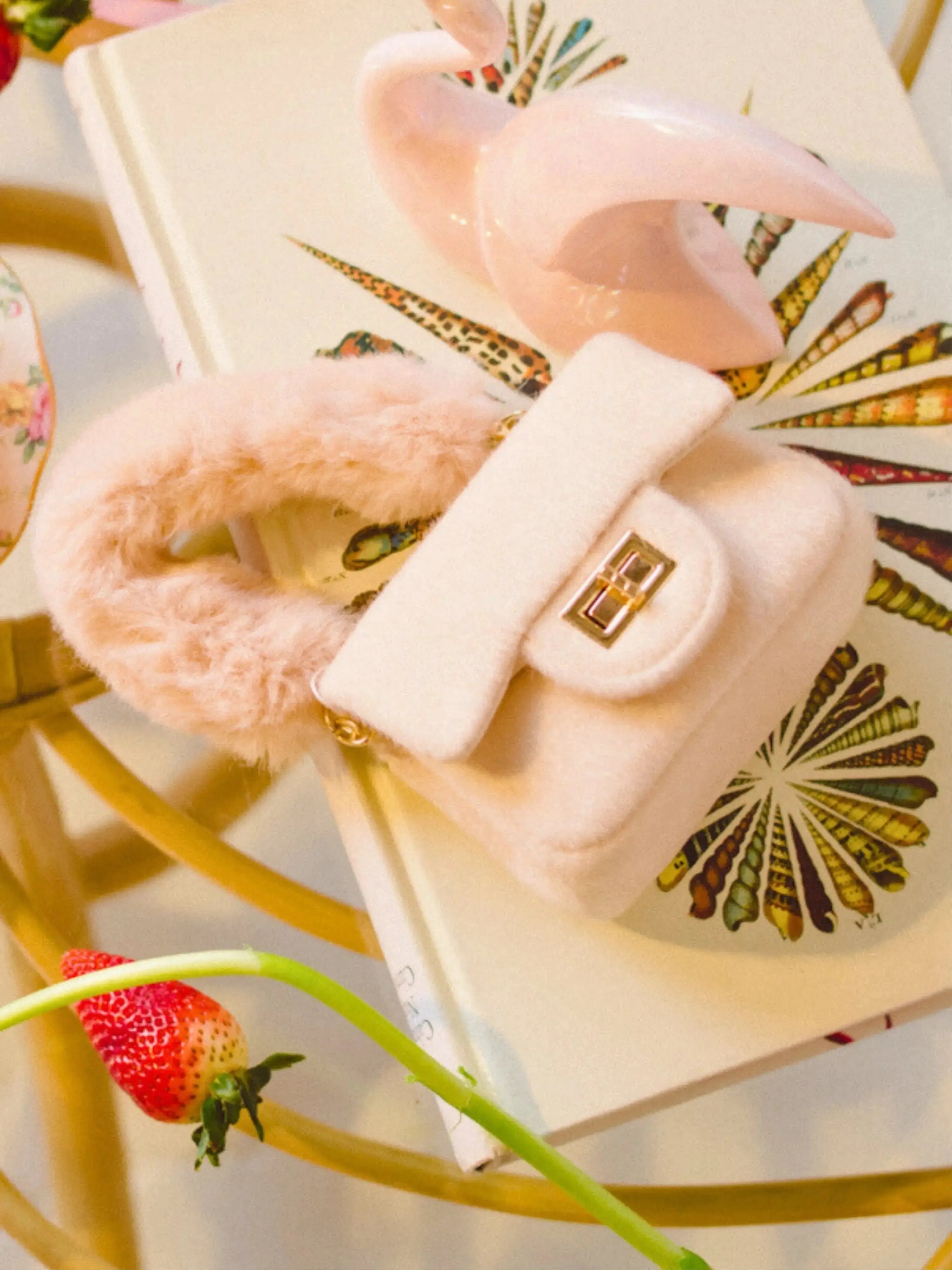 The Petit Agneau Tiny Purse in ivory with faux fur handle and gold clasp, styled on a decorative book for chic micro handbag appeal.